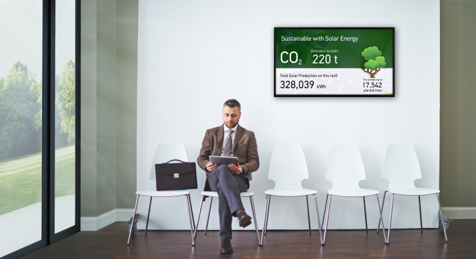 a man ona laptop sits in a waiting roof in front solar a Solarfox display screen which is presenting the solar data from the buildings solar PV system